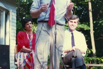 Man speaking into microphone outdoors with two seated listeners.