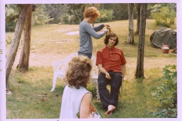 A person styles another's hair outdoors while a third watches.