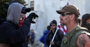 A man in a uniform talks to a masked protester holding a sign.